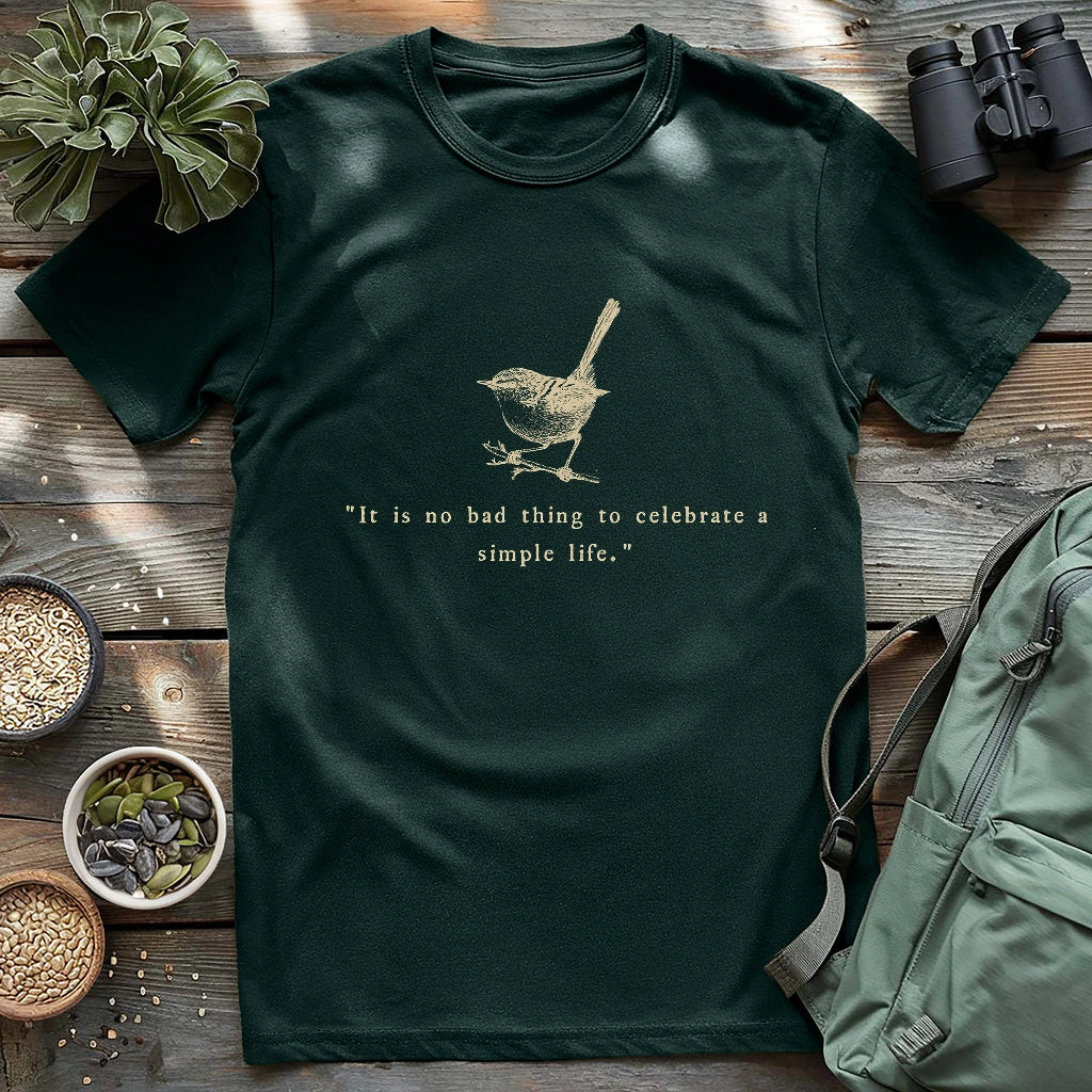 Forest green t-shirt featuring a beige sketch of a wren perched on a branch above the quote “It is no bad thing to celebrate a simple life,” styled flat with nature-themed props.