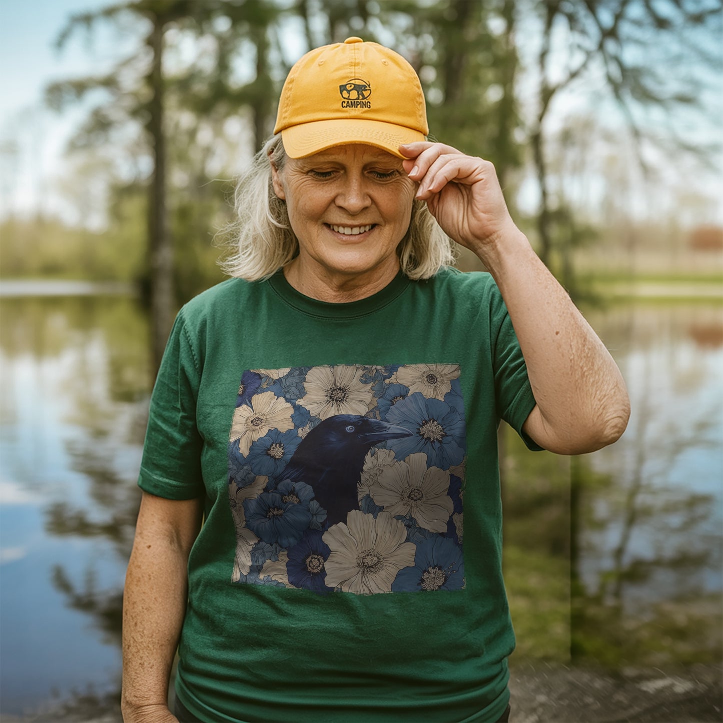 Person wearing a forest green t-shirt with raven in flower field design and yellow cap outdoors by a lake