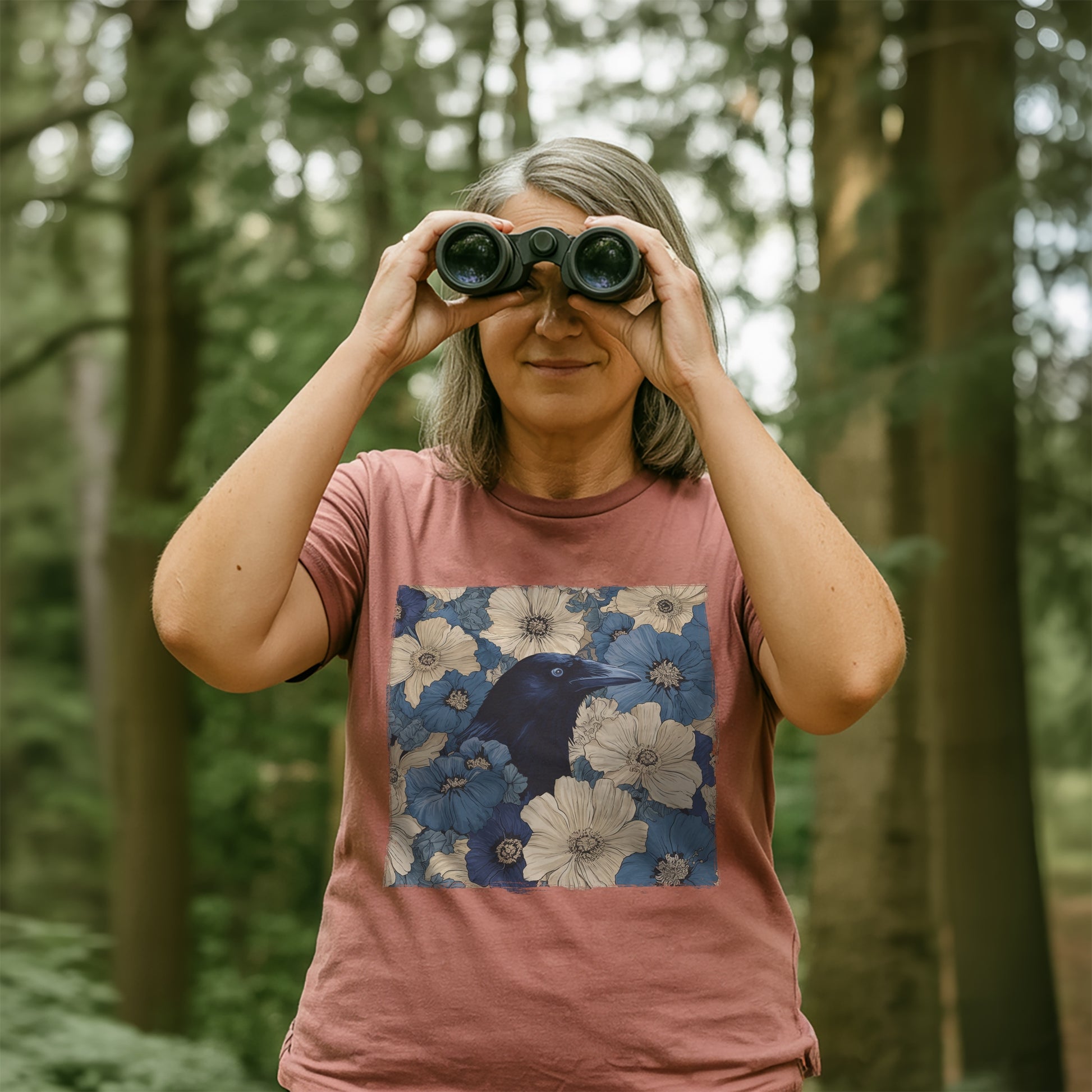 Women in a forest using binoculars, wearing a heather mauve t-shirt with a raven in flower field design.