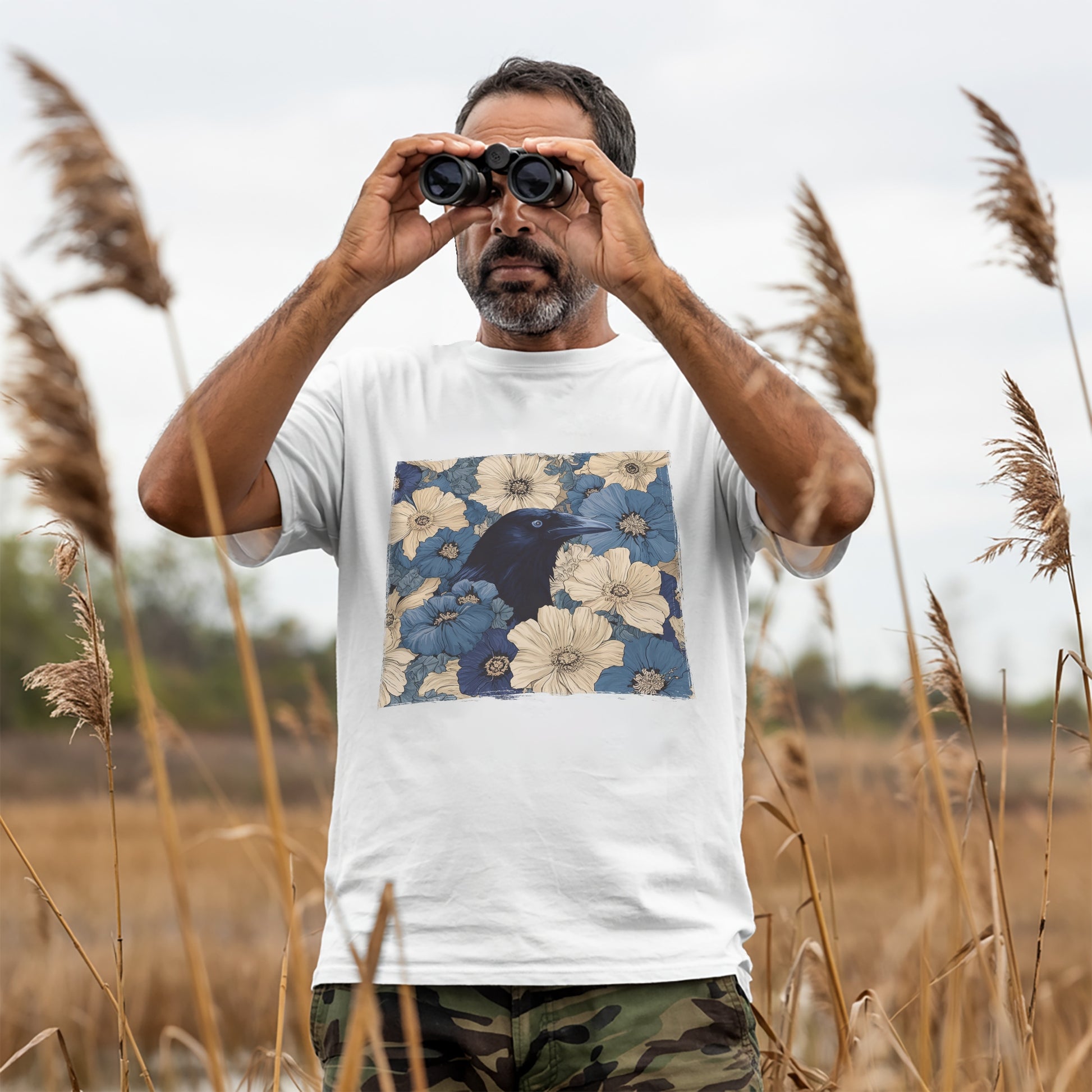 Man in a field using binoculars, wearing a white t-shirt with a raven in flower field design.