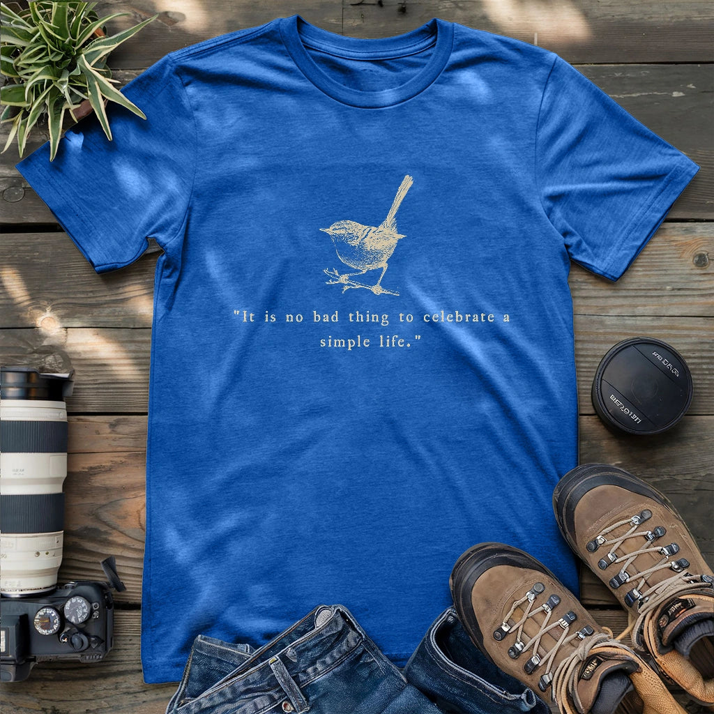 Royal blue t-shirt featuring a beige hand-drawn wren and the quote “It is no bad thing to celebrate a simple life,” styled flat with hiking boots, jeans, and camera gear.