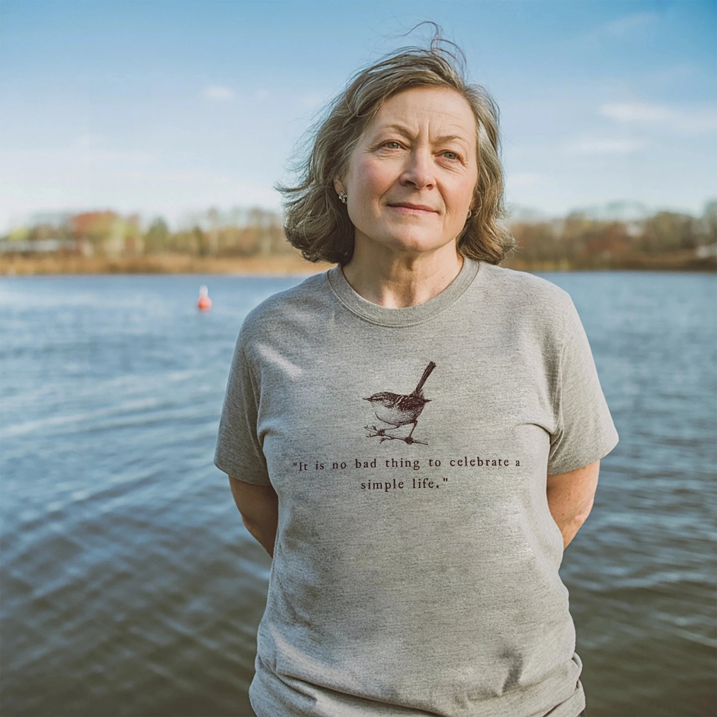 Woman standing peacefully by a lake wearing a athletic heather t-shirt featuring a hand-drawn bird and the quote “It is no bad thing to celebrate a simple life,” with trees and blue sky in the background.