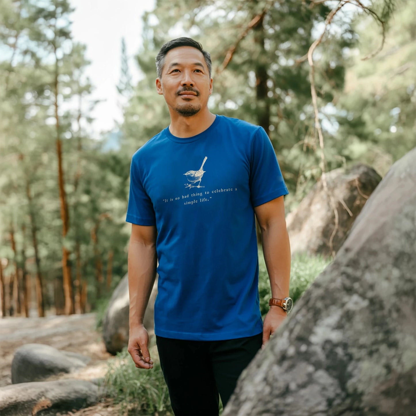 Man walking along a rocky forest trail wearing a royal blue t-shirt with a sketched bird and the quote “It is no bad thing to celebrate a simple life,” surrounded by tall pine trees.
