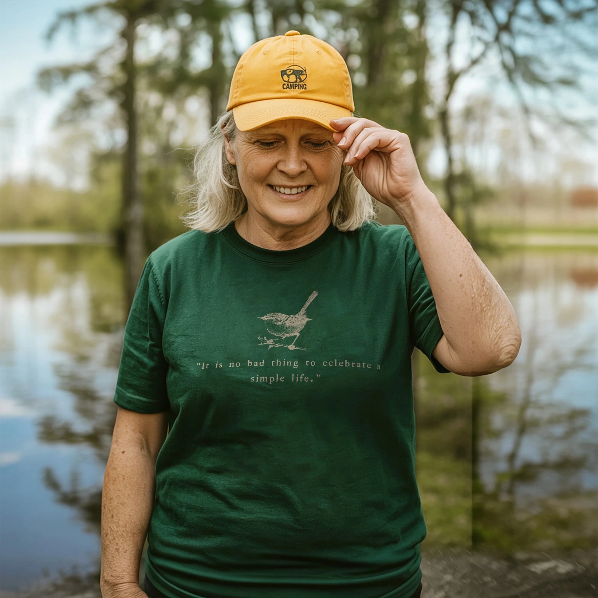 Smiling woman wearing a forest green t-shirt with a vintage bird illustration and the quote “It is no bad thing to celebrate a simple life,” along with a yellow camping hat, standing by a calm lake surrounded by trees.