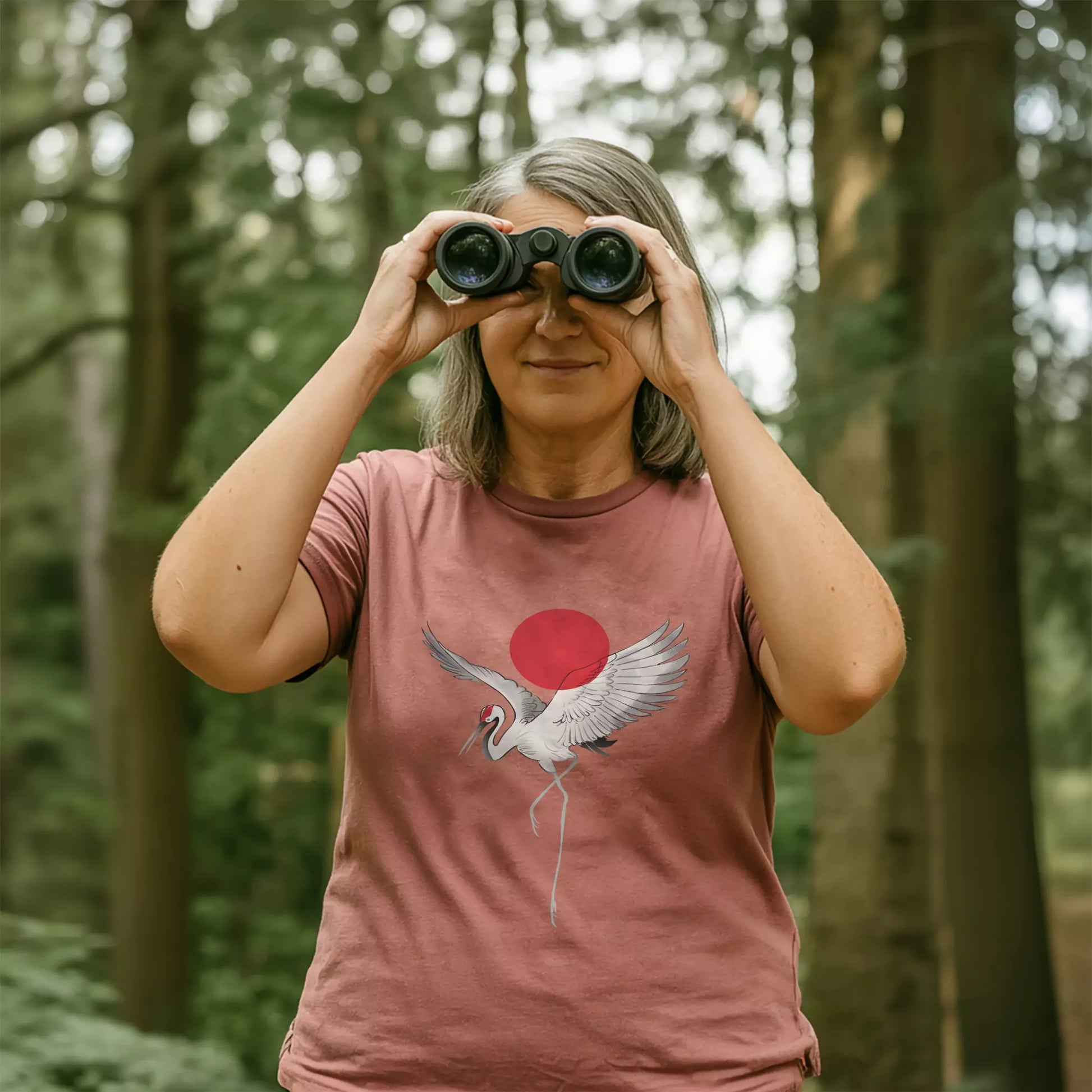 Woman birdwatching in the woods wearing a heather mauve t-shirt with a white crane and red sun print, holding binoculars in a lush forest setting.
