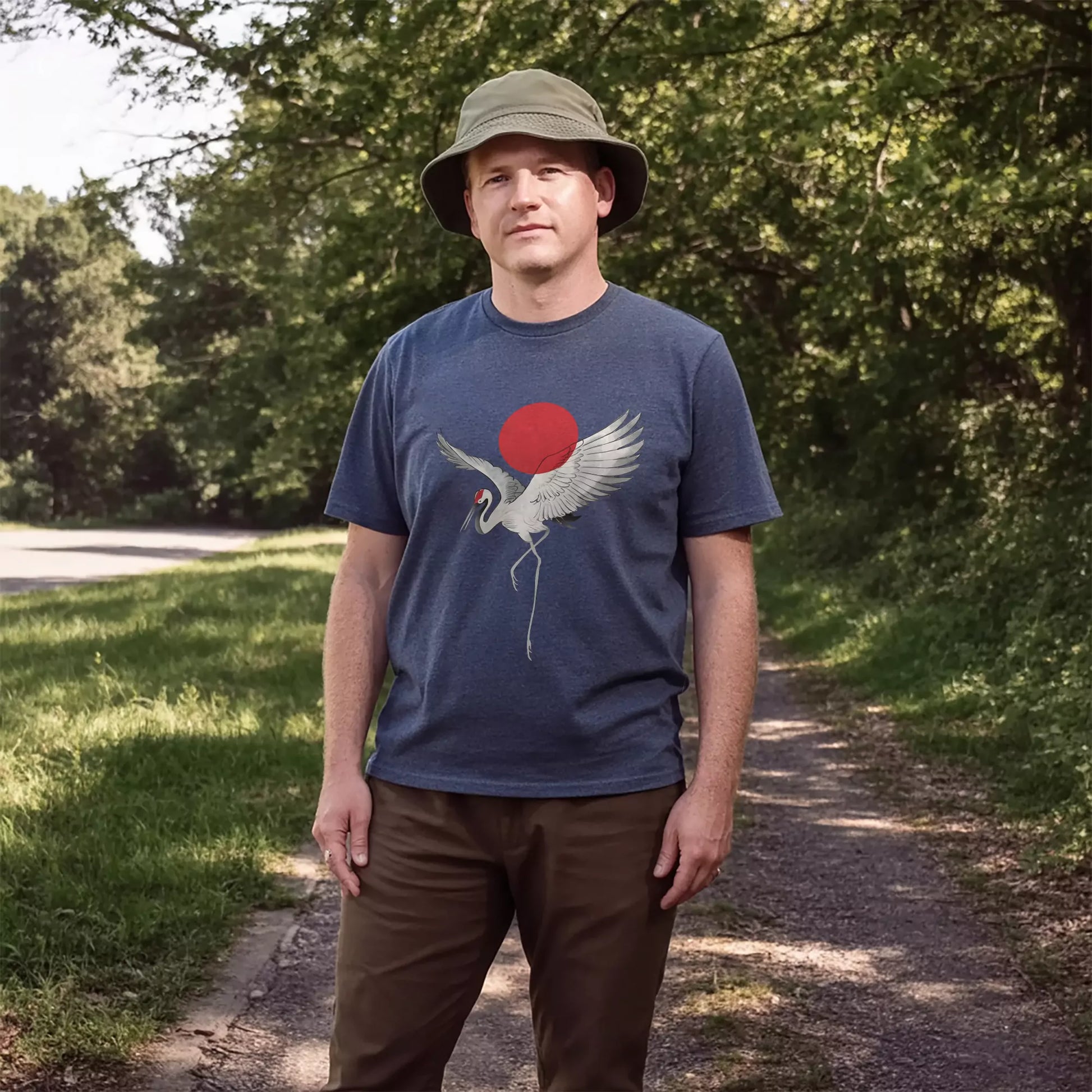 Man wearing a heather navy t-shirt featuring a white crane and rising red sun, standing on a forested trail with relaxed, outdoor styling.