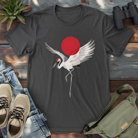 Dark grey heather t-shirt featuring a white crane with outstretched wings flying in front of a red sun, styled with casual sneakers, jeans, and a green sling bag on a wooden surface.