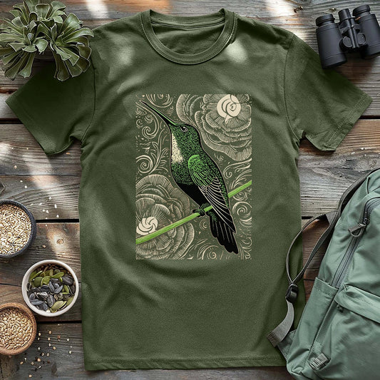 Military green t-shirt featuring a vibrant green hummingbird perched on a branch with swirling floral patterns in the background, styled flat with outdoor accessories.