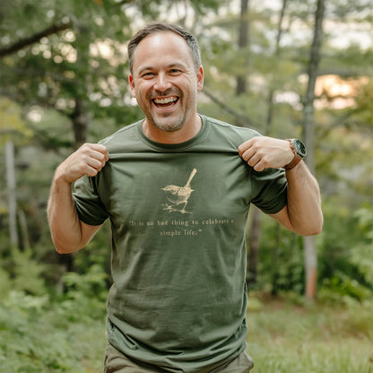 Smiling man holding out his olive green t-shirt featuring a sketched bird and the quote “It is no bad thing to celebrate a simple life,” standing in a sunlit forest setting.